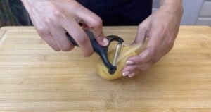 Person peeling a potato with a peeler on a wooden cutting board, showcasing food preparation techniques.